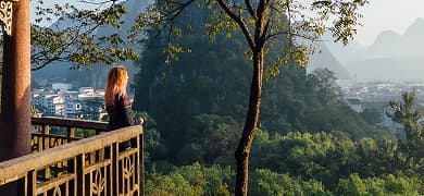 A woman stands on a balcony overlooking a jungle. A tall tree stands in front of her.