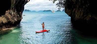 A woman stands on a paddleboard on the water. She is positioned between two stoney outcroppings with greenery.
